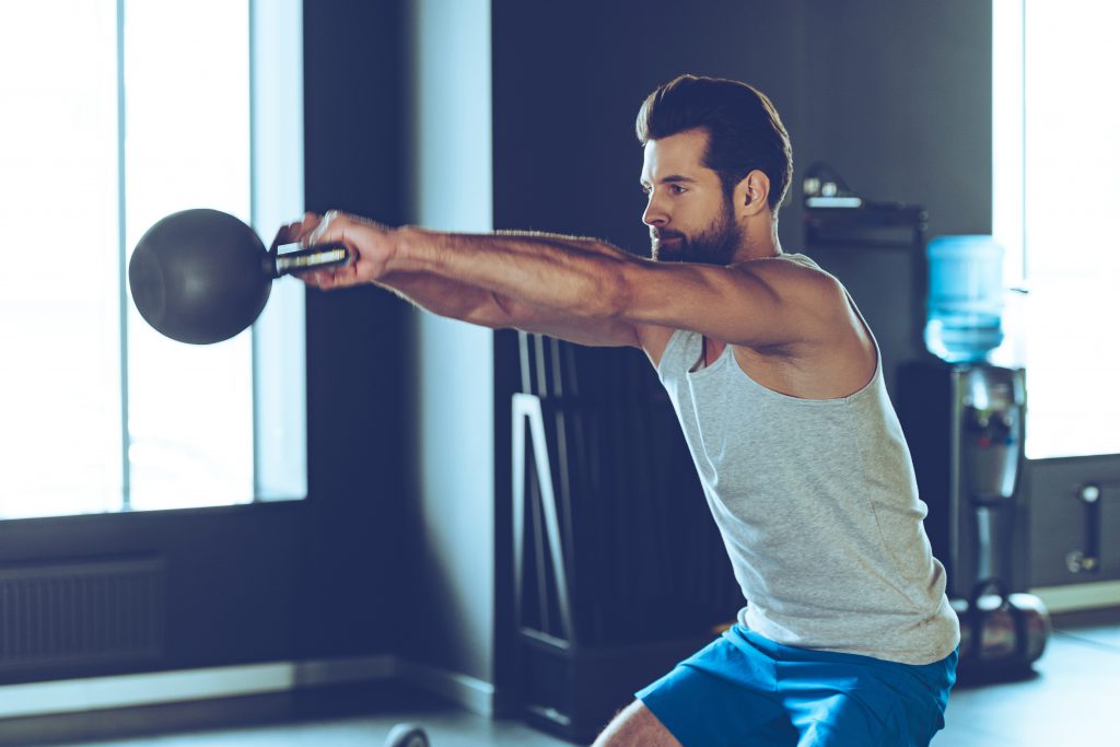 Side view of young handsome man in sportswear working out with kettle bell at gym